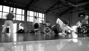 Immagine chee ritrae danzatori e danzatrici con disabilità e non, impegnati nel movimento e nella danza, in uno spazio circoscritto; si vedono carrozzine e persone in pedi. L'immagine restituisce velocità e dinamicità.